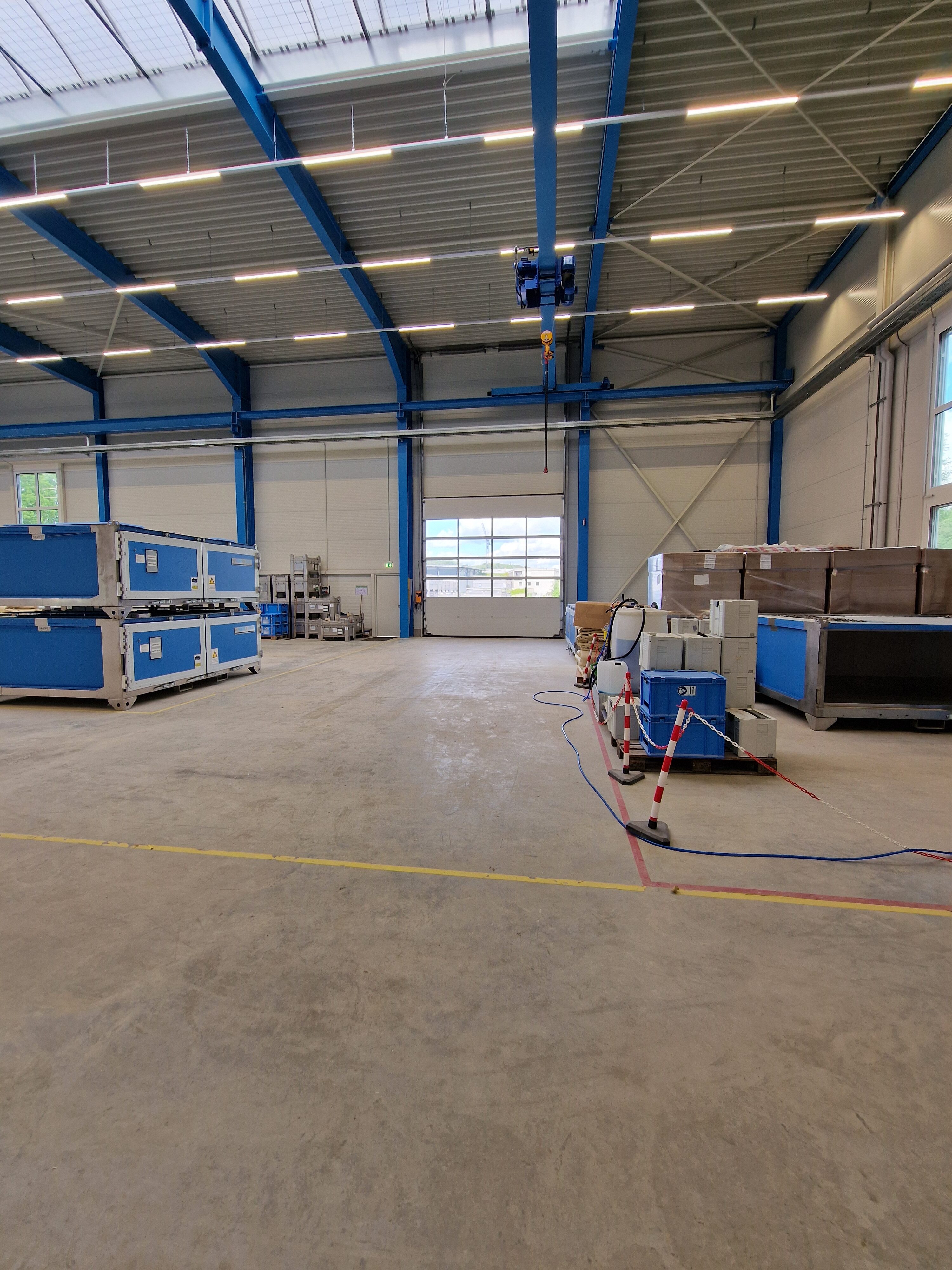 Eine leere Lagerhalle mit blauen Kisten und hellem Betonboden; große Fenster und Deckenbeleuchtung.