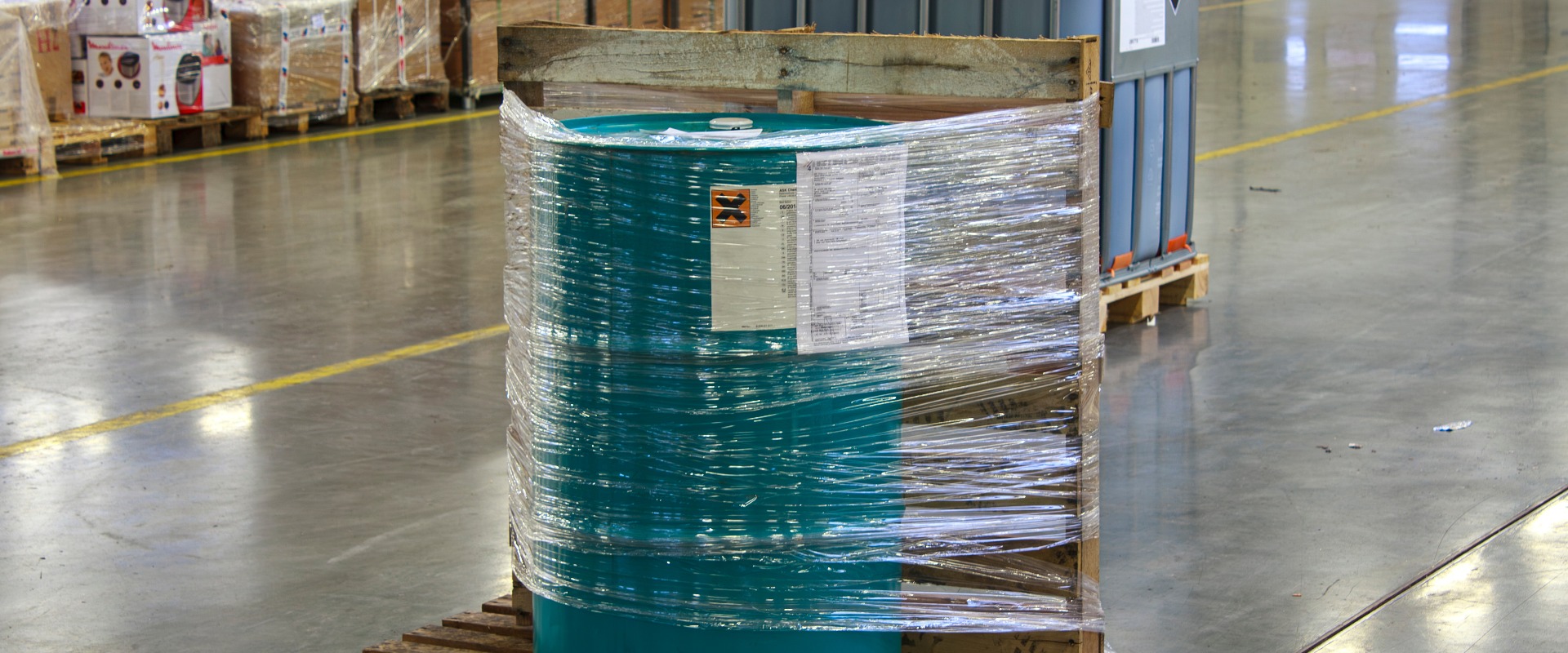 Grünes Fass, das in Plastik eingewickelt und auf einer Holzpalette befestigt ist. Das Fass ist von verpackten Waren und einem weiteren großen Container umgeben, was auf eine organisierte Lagerumgebung hindeutet.